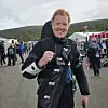 Person i svart kjøredrakt smiler foran et pitområde med folk, biler og flagg.
