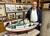 En mann står bak en stor modellbåt i et rom med maritime fotografier og utstilte objekter på veggene.