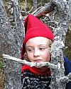 Barn med rød nisselue kikker mellom trestammer i skogen.