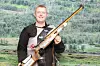 En mann står med en rifle mot en bakgrunn av grønn skog og åpent terreng.