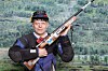 En mann i skytterutstyr holder en rifle foran en bakgrunn med skogkledd fjellandskap.