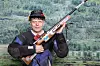 En mann i skytterutstyr holder en rifle foran en bakgrunn med skogkledd fjellandskap.