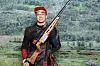 Mann i skytterutstyr holder en rifle foran en bakgrunn med grønt fjellandskap.