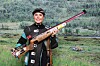Dame i skytterutstyr holder en rifle foran en bakgrunn med grønt skoglandskap.