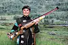 Dame i skytterutstyr holder en rifle foran en bakgrunn med grønt skoglandskap.