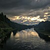 Stille elv med speilblankt vann under mørke skyer og glimt av solnedgang mellom skogkledde bredder.