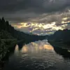 Stille elv med speilblankt vann under mørke skyer og glimt av solnedgang mellom skogkledde bredder.