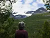 Person sett bakfra foran en grønn dal med fjell og snøfelt i landskapet.