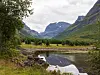 Grønn fjelldal med elv, vannspeil og bratte fjell under en skyet himmel.