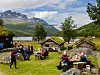 Folk sitter og står ved bord i et grønt fjellområde med hytter, innsjø og snødekte fjell i bakgrunnen.