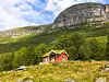 To røde hytter står på en gresskledd åsside foran et stort fjell med skog og stein.