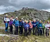 En gruppe fotturister står på et steinete terreng foran en fjellrygg under overskyet himmel.