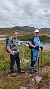 To turvandrere står ved et skilt i et fjellandskap med vann og lave fjell i bakgrunnen.