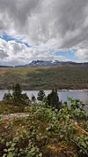 Skyet himmel over fjell, innsjø og skogkledd forgrunn.