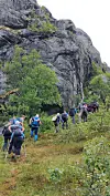En gruppe vandrere går på sti ved en bratt fjellvegg med busker og trær.