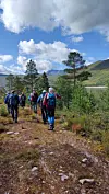 Turgåere med sekk går på en steinete sti i fjellterreng med trær og innsjø.