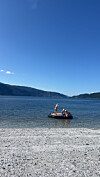 Flere personer står og sitter på en badeflåte ved en steinstrand med fjell og åpent vann i bakgrunnen.