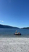 Flere personer står og sitter på en badeflåte ved en steinstrand med fjell og åpent vann i bakgrunnen.