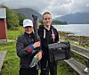 To personer står utendørs ved sjøen med fiskeutstyr og en svart bag.