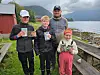 Tre barn står utendørs ved fjorden og holder drikker, med fjell og overskyet himmel bak.