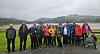 En gruppe personer står på en grusplass foran grønt landskap, flere med turstaver, og en rullestol står til høyre.