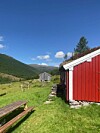 Rød hytte ved en gresskledd sti med fjell og blå himmel i bakgrunnen.