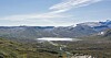 Vid utsikt over grønn dal, snødekte fjell og en smal elv som renner gjennom landskapet.