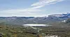 Vid utsikt over grønn dal, snødekte fjell og en smal elv som renner gjennom landskapet.