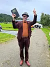 Mann i brun dress og hatt holder en bok og peker oppover på en grusvei.