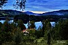 Utsikt over innsjø omgitt av skog og åser med en liten bygning i forgrunnen ved solnedgang.