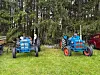 To blå traktorer står på gresset foran tett skog, med en rød bil delvis synlig bak.