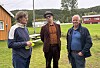 Tre personer står ute på gras ved et idrettsanlegg med tribune og mål i bakgrunnen.