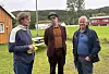 Tre personer står ute på gras ved et idrettsanlegg med tribune og mål i bakgrunnen.