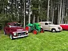 Klassiske biler og en grønn traktor står parkert på en gressplen ved skog.