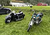 To veteranmotorsykler står på en gresslette foran bygninger i et landlig område.