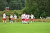 Gruppe fotballspillere i hvite og røde drakter på en gressbane foran skog og små bygninger.