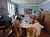 En gruppe mennesker sitter rundt et langbord i en lys, tradisjonell spisestue med antikke møbler og trofeer.