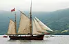 Et historisk seilskip med høye master og hvite seil på en fjord med fjell i bakgrunnen.