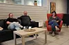 Tre personer sitter i et loungeområde med sofaer, bord og røde stoler i et lyst innemiljø.