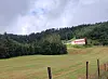 Hvitt gårdshus med rødt tak i grønt jordbrukslandskap ved en skogkledd ås.