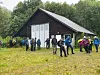 Gruppe turgåere står foran en mørk trehytte i et vått gressområde ved skog.