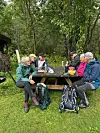 Fem turgåere sitter ved et bord på en skogkledd rasteplass med sekker og drikke.
