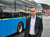 En mann i mørk jakke står ved siden av en blå buss utendørs.