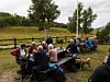Folk sitter rundt langbord utendørs i en park med grønne trær og en hund i forgrunnen.