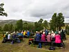 Gruppe mennesker samlet ved piknikbord i et grønt landskap med fjell i bakgrunnen.