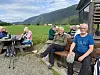 Personer sitter på benker og ved et bord utenfor en hytte med fjellandskap i bakgrunnen.