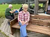 Kvinne sitter på en trebenk foran et lite hus, med en mann  i bakgrunnen.