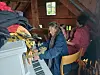 To personer sitter ved et piano i et loftsrom med stablede klær og esker rundt seg.