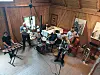 Et band øver i et lyst loftsrom med trevegger, trommer, keyboard og blåseinstrumenter.