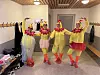 Fire personer i fargerike kostymer poserer i et garderoberom med benker og knagger.